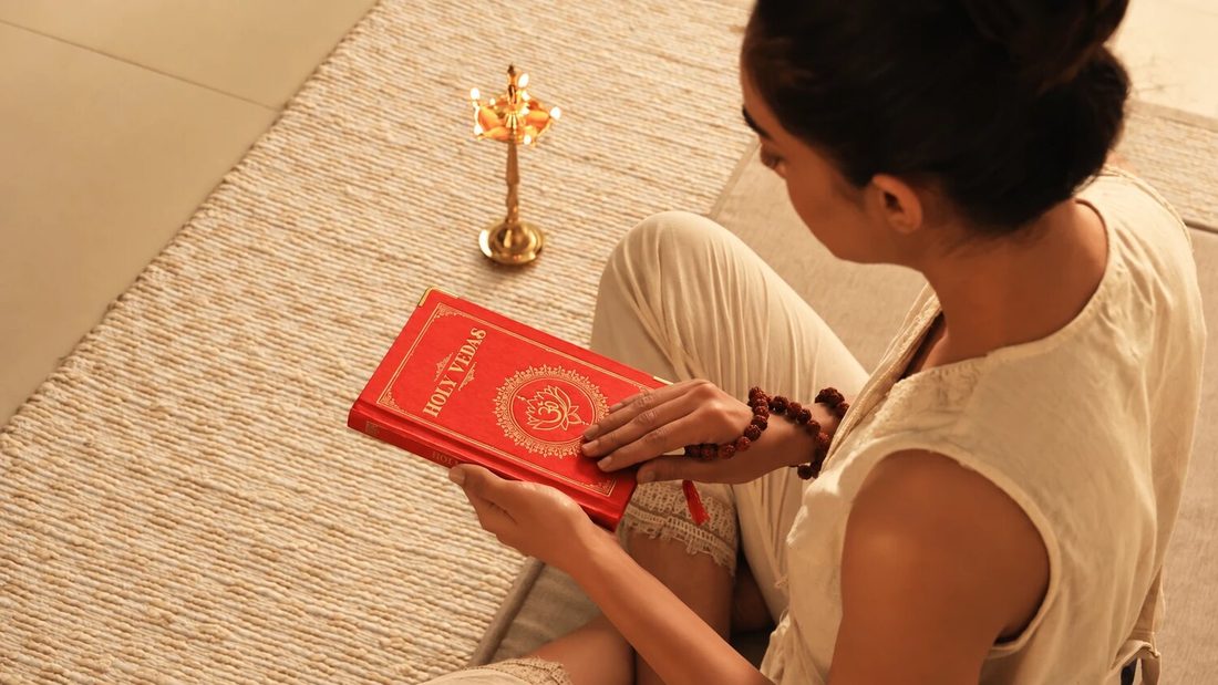 Art of Vedas: kobieta siedząca na plecionej macie czyta czerwoną księgę Świętych Wed obok mosiężnej lampy oliwnej